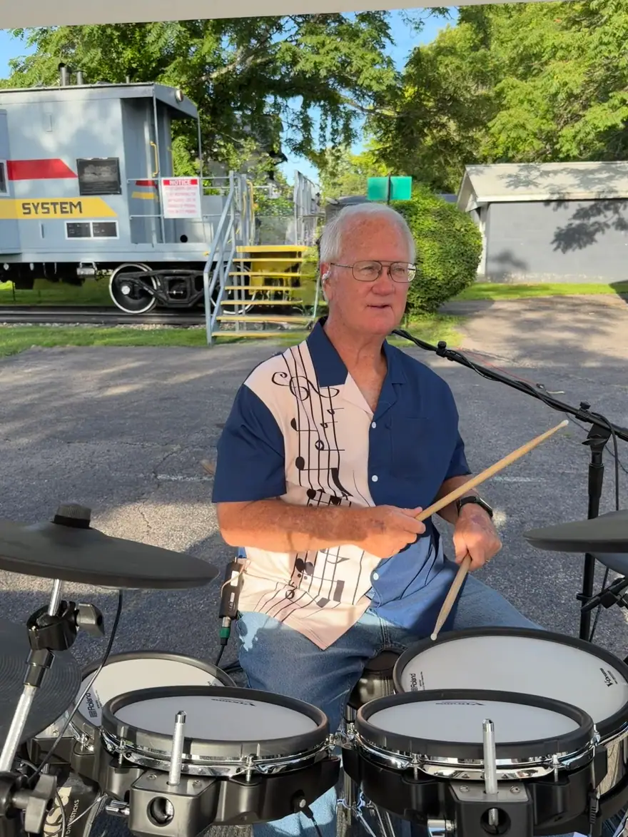 Ventura Band drummer in a navy music-note shirt playing beside a red train caboose