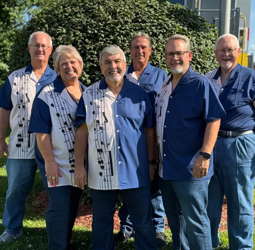 Six members of The Ventura Band in navy music-note shirts, standing together outdoors by a hedge