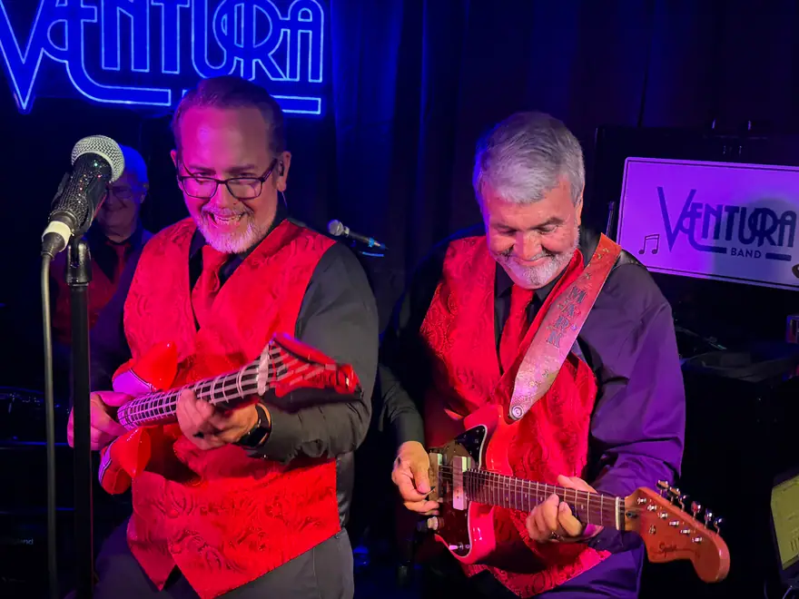 Two Ventura Band guitarists in red paisley vests playing under a neon Ventura sign