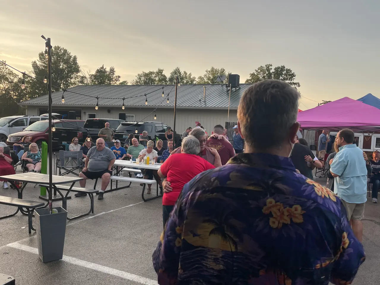 People gathered outdoors at dusk around picnic tables, a man in a Hawaiian shirt in front